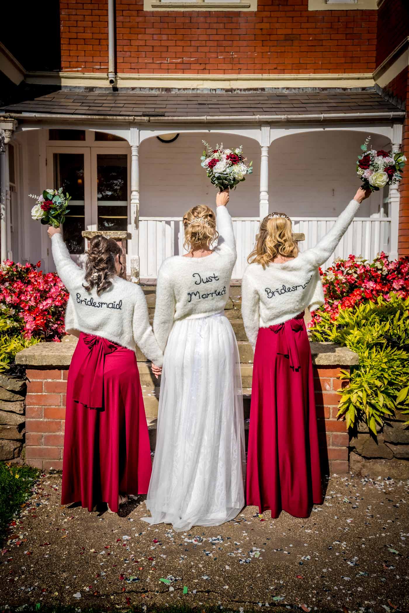 Bride with two bridesmaids in red from the back holding bouquets in air and with writing on their backs