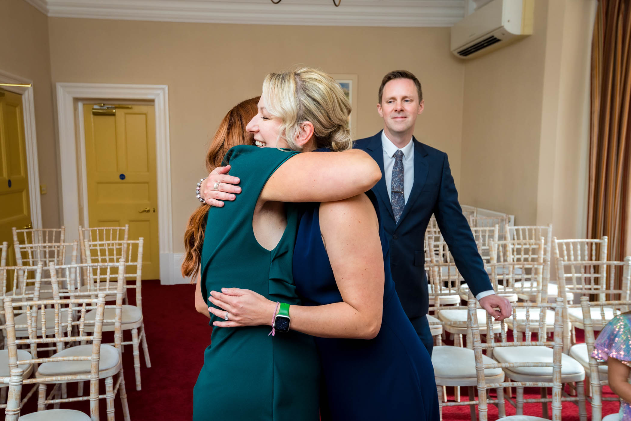 Two ladies hug at wedding whilst man watches
