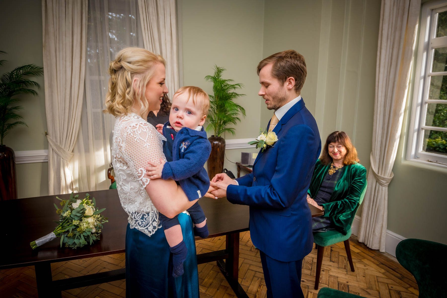 Groom places ring on the finger of his bride who holds their toddler son during wedding at Richmond Register Office.