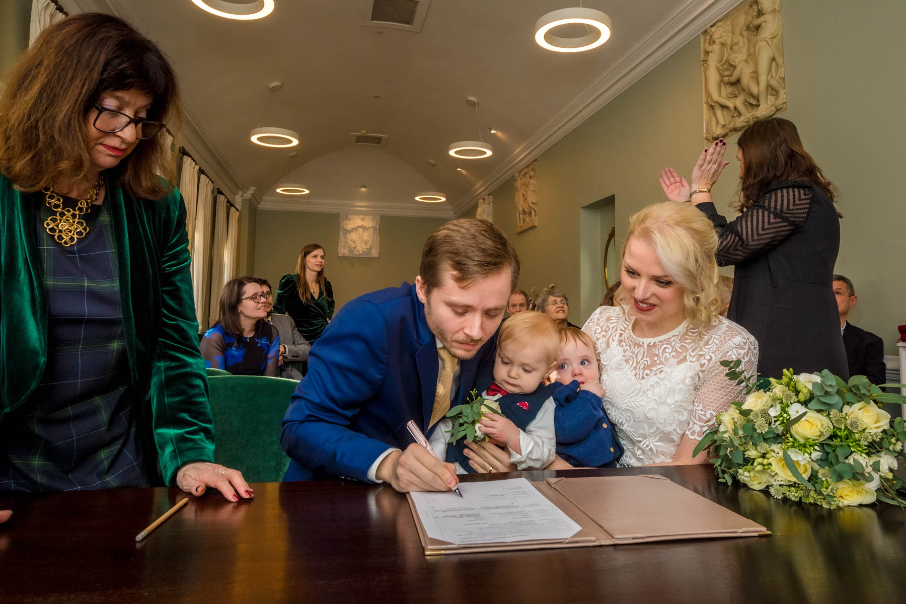 Groom signs wedding schedule as bride and 2 children watch in Loggia Room at York House.