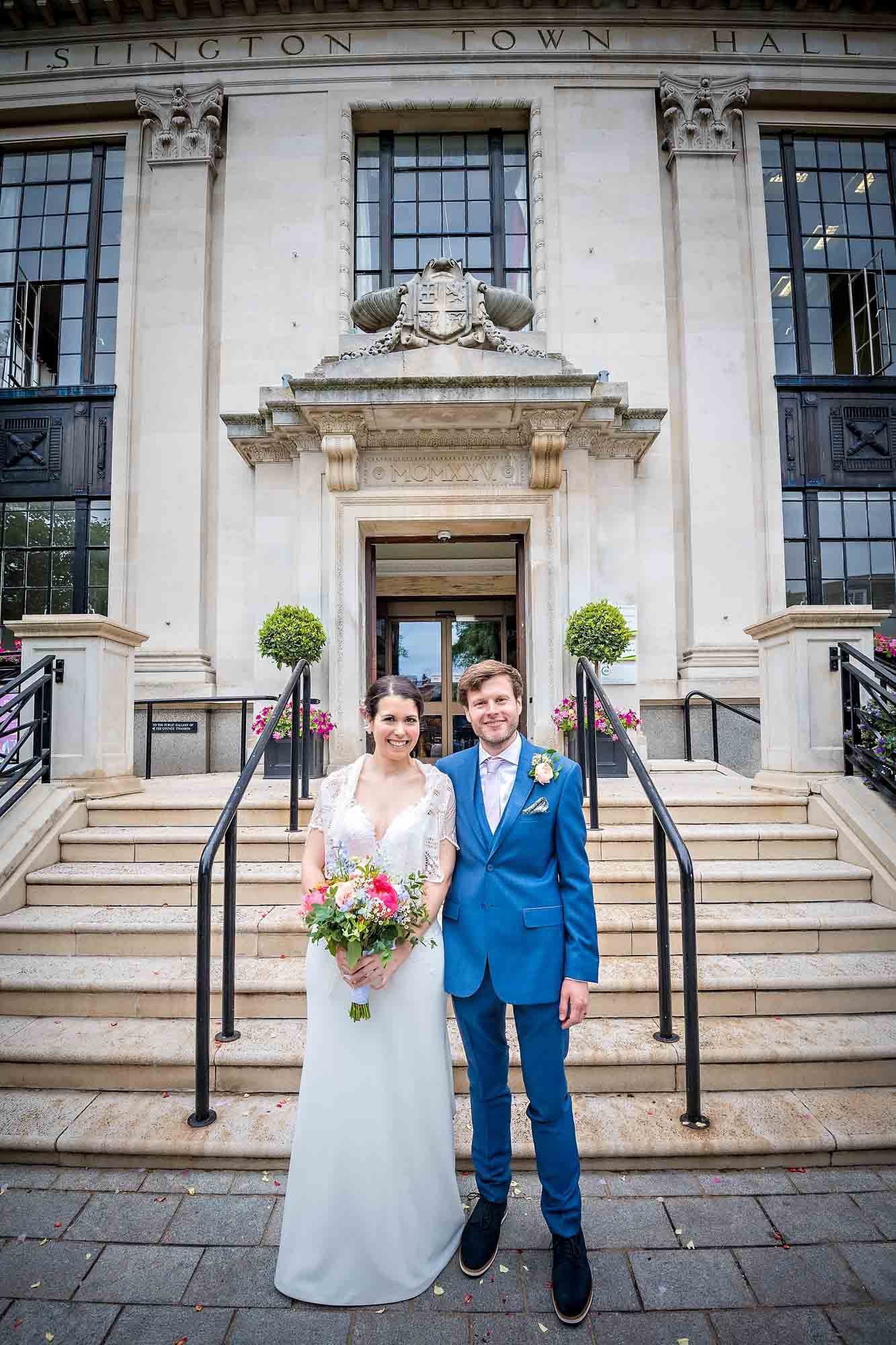 Bride with bouquet and groom in blue suit pose in front of Islingtom Town Hall