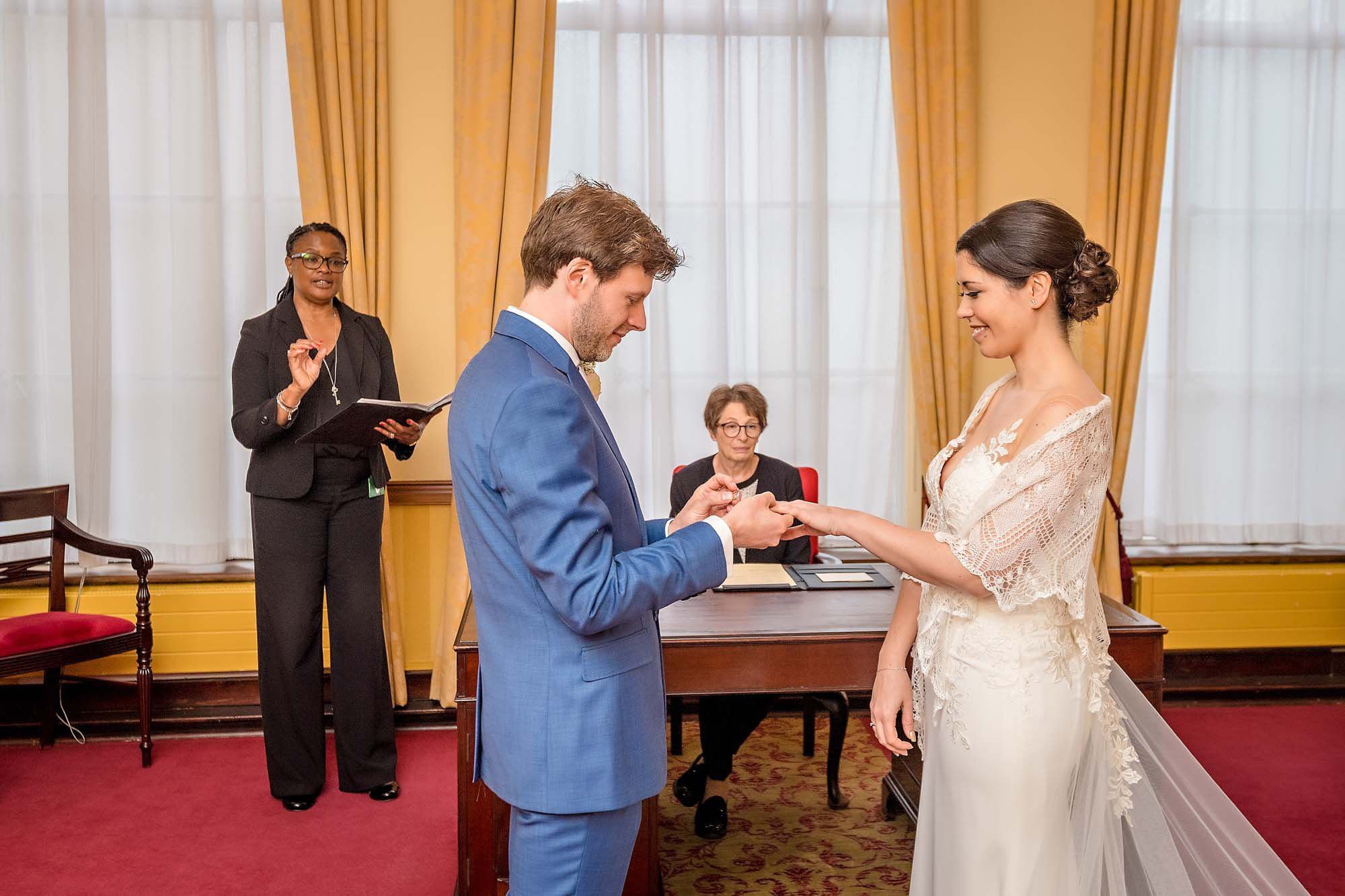 In Room 99 at Islington Town Hall, the groom proudly places the ring on his bride's finger as she smiles