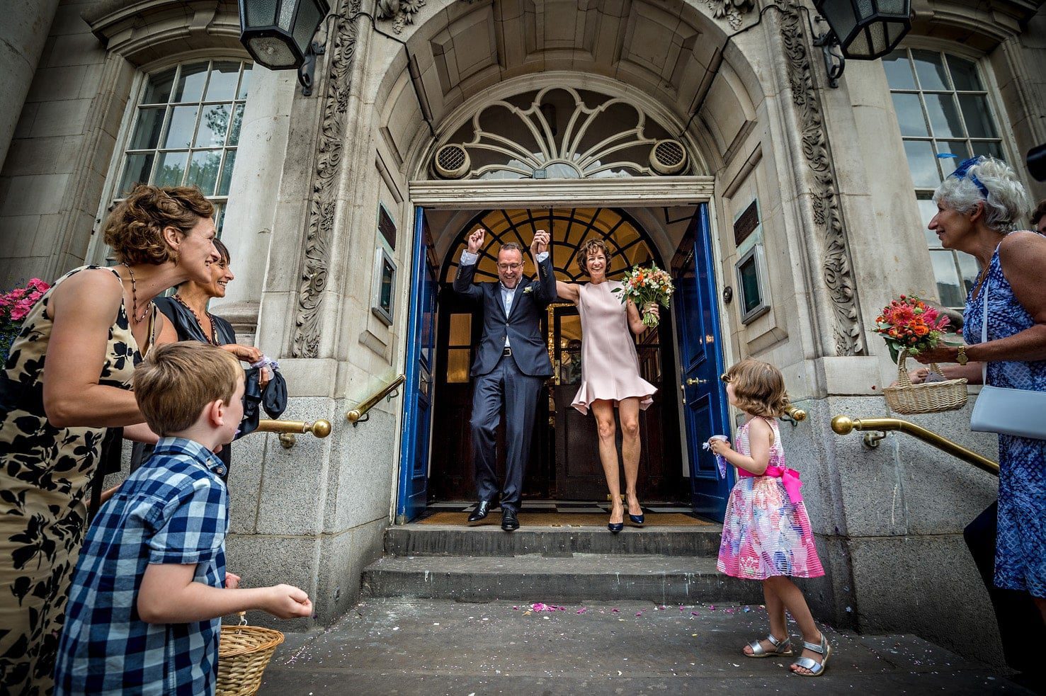 Newlyweds holding hands above their heads and rushing out of Chelsea Old Town Hall to greet their guests after their short wedding