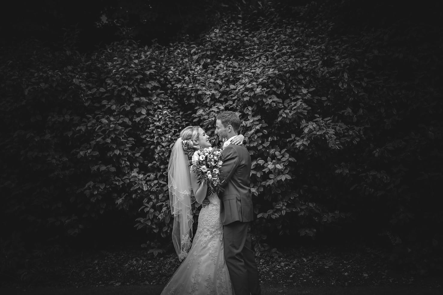 Wedding Portrait of Groom Holding Bride with Bouquet In Front of Bushes in Black and White