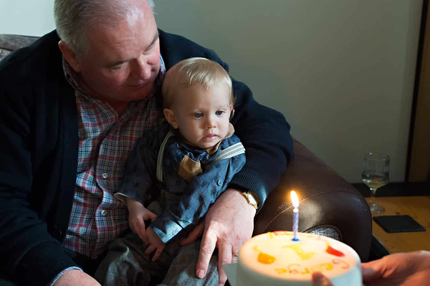 Toddler sitting on granddad's lap looking at birthday cake in Caerphilly