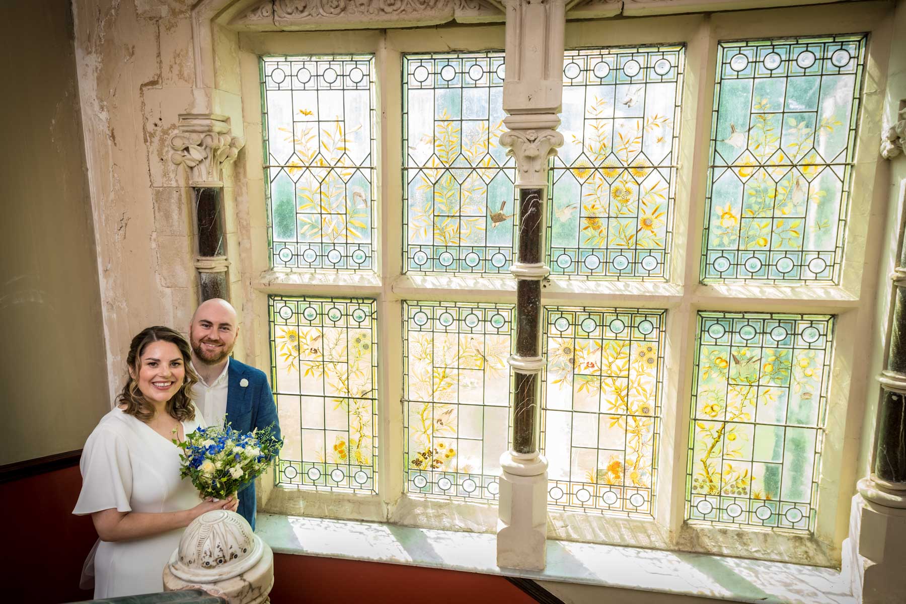 Newlyweds posing with large stained glass window on the landing of Insole Court