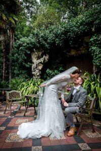 Bride kissing seated groom in garden seating area at Hampton Court House
