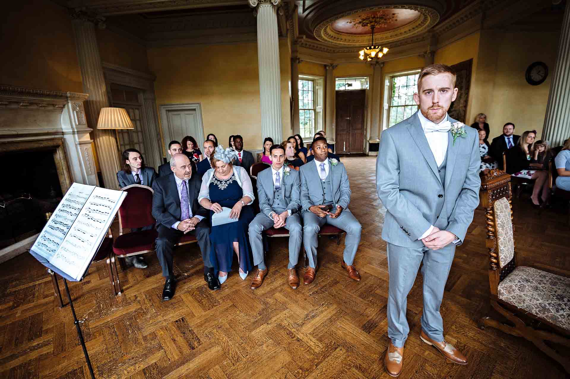 Groom looking serious with guests in background in Grand Hall of Hampton Court House
