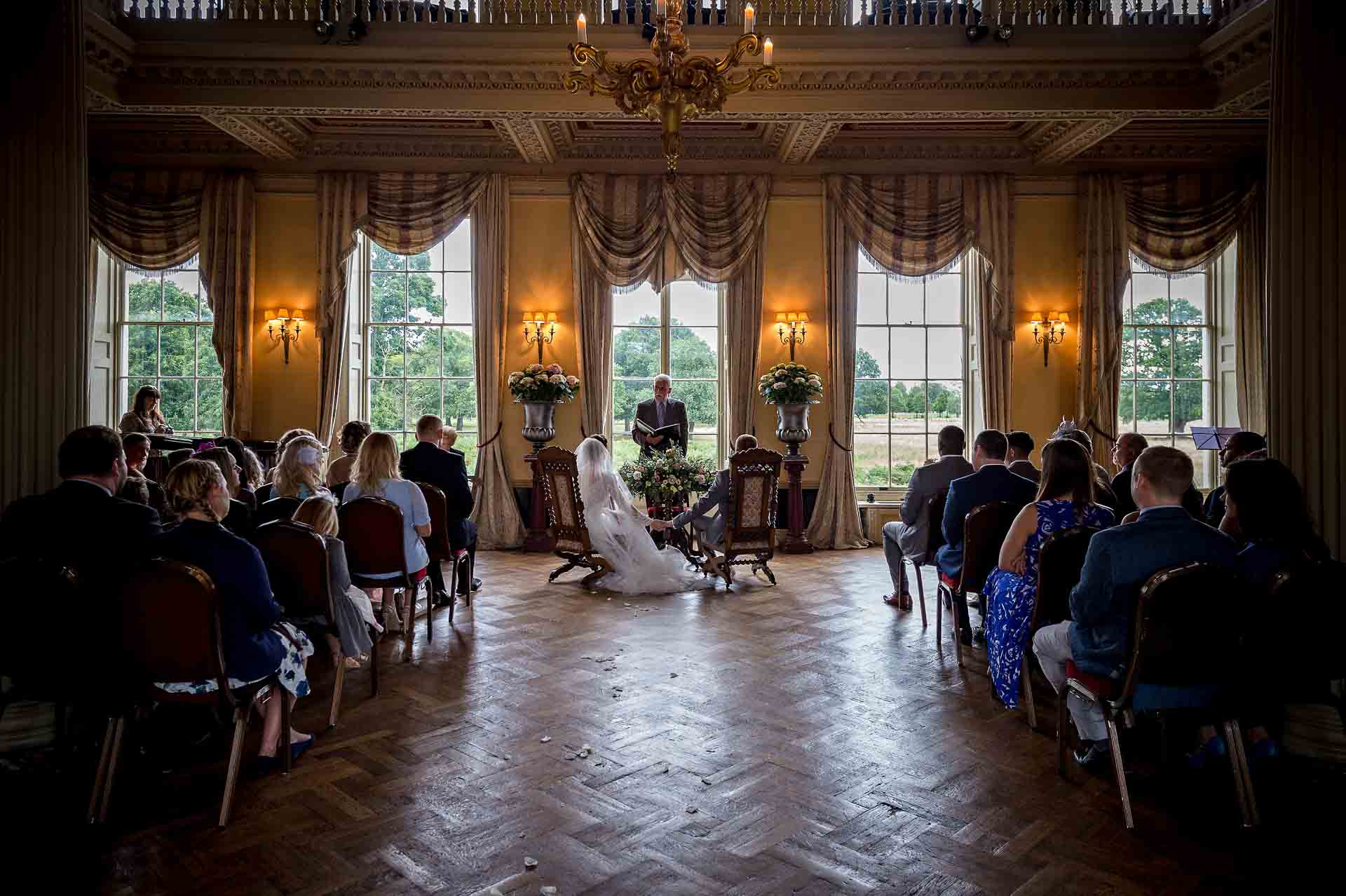 Shot from back of Hampton Court House wedding as seated bride and groom hold hands