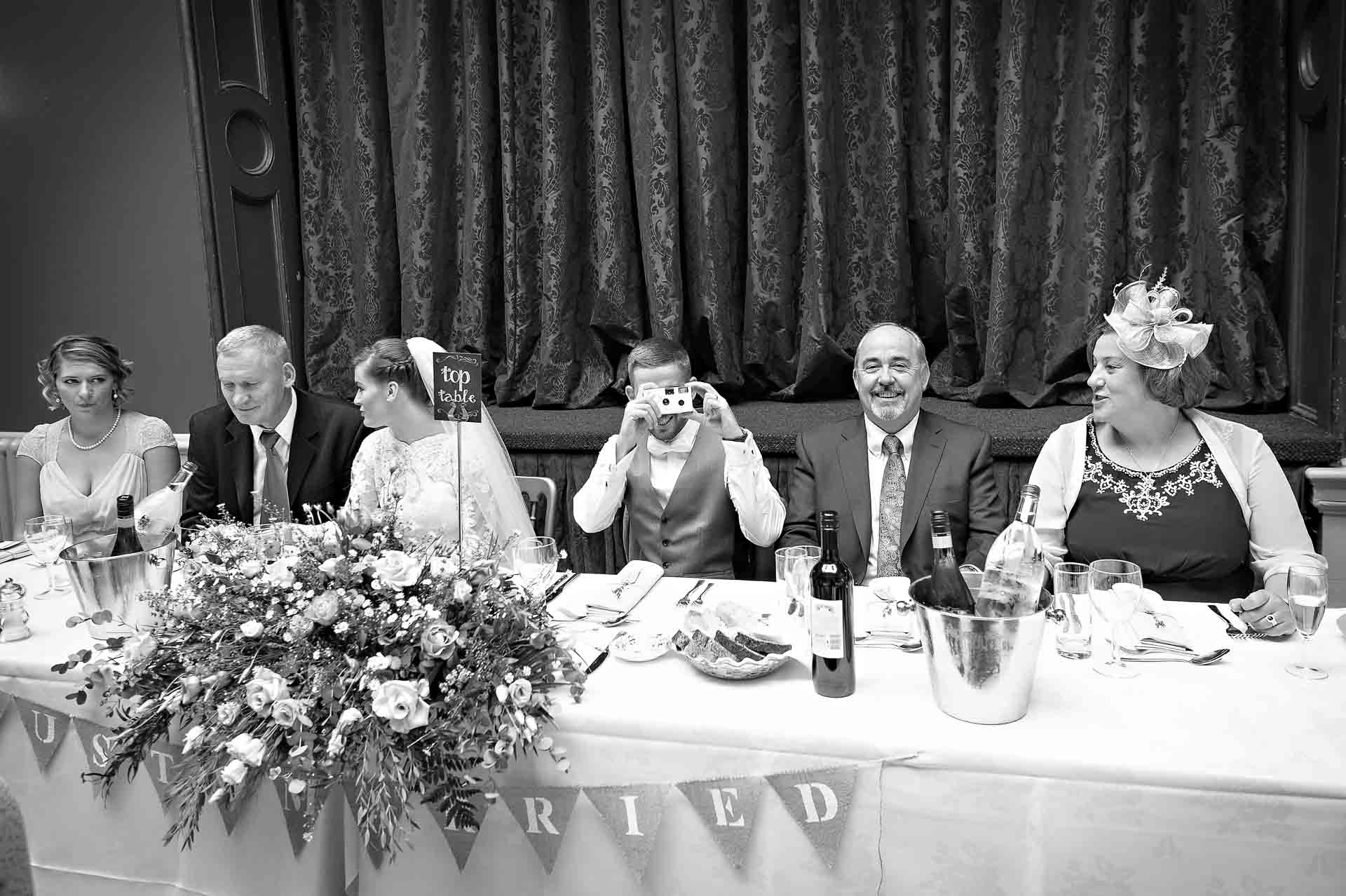 Groom taking photo of the photographer whilst other guests talk at top table at wedding reception