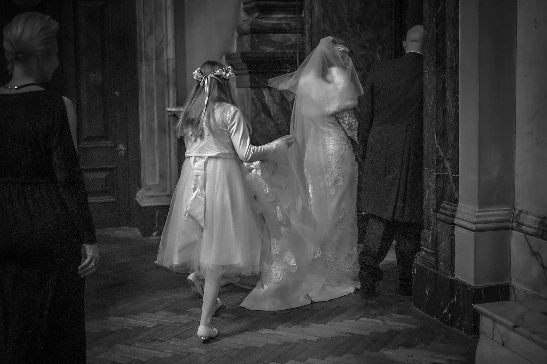 A bridesmaid holds the brides dress as they process through to the sacristy for the signing of the schedule at Brompton Oratory