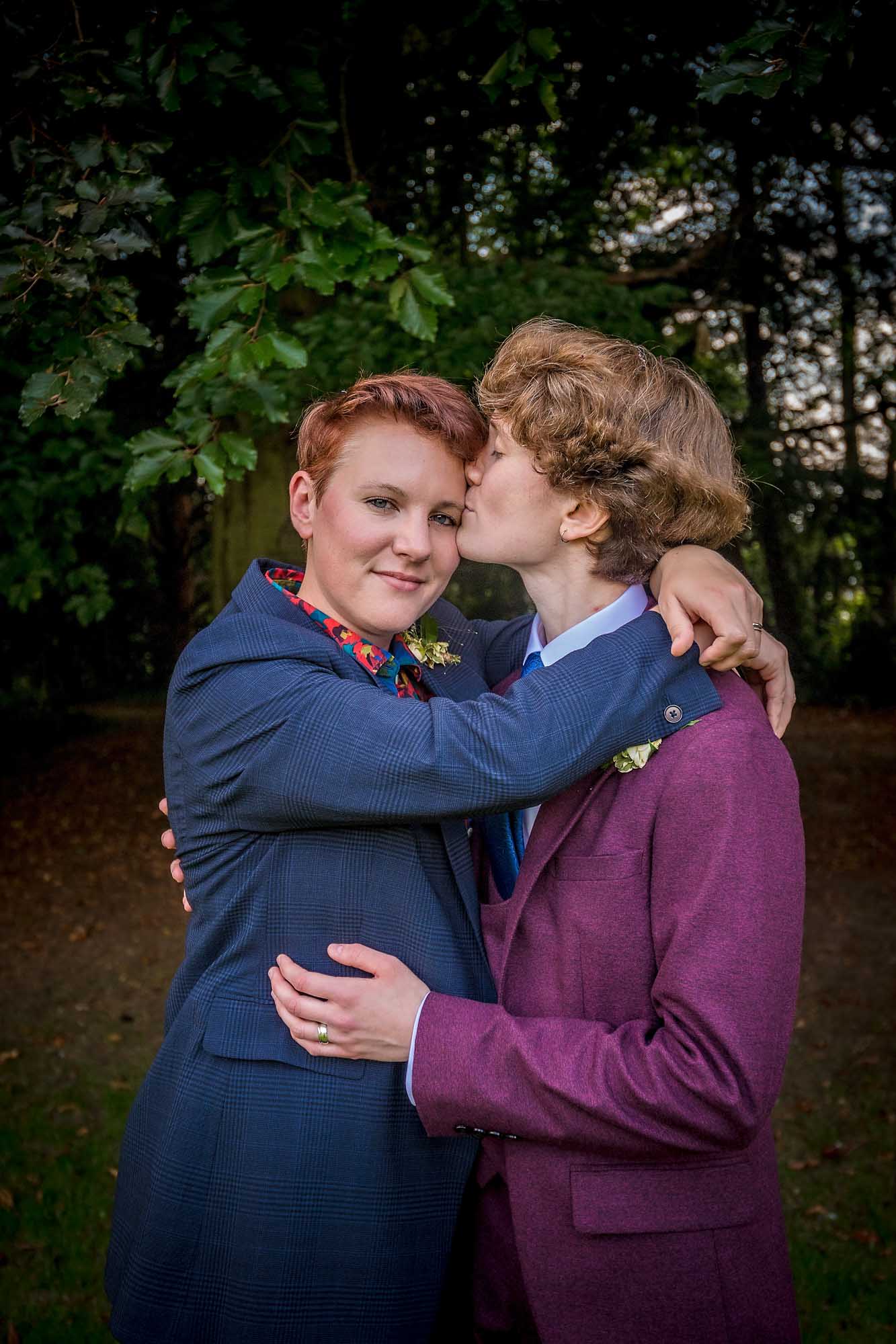 Bride gives camera eye-contact whilst her bride kisses her on cheek at LGBTQ wedding