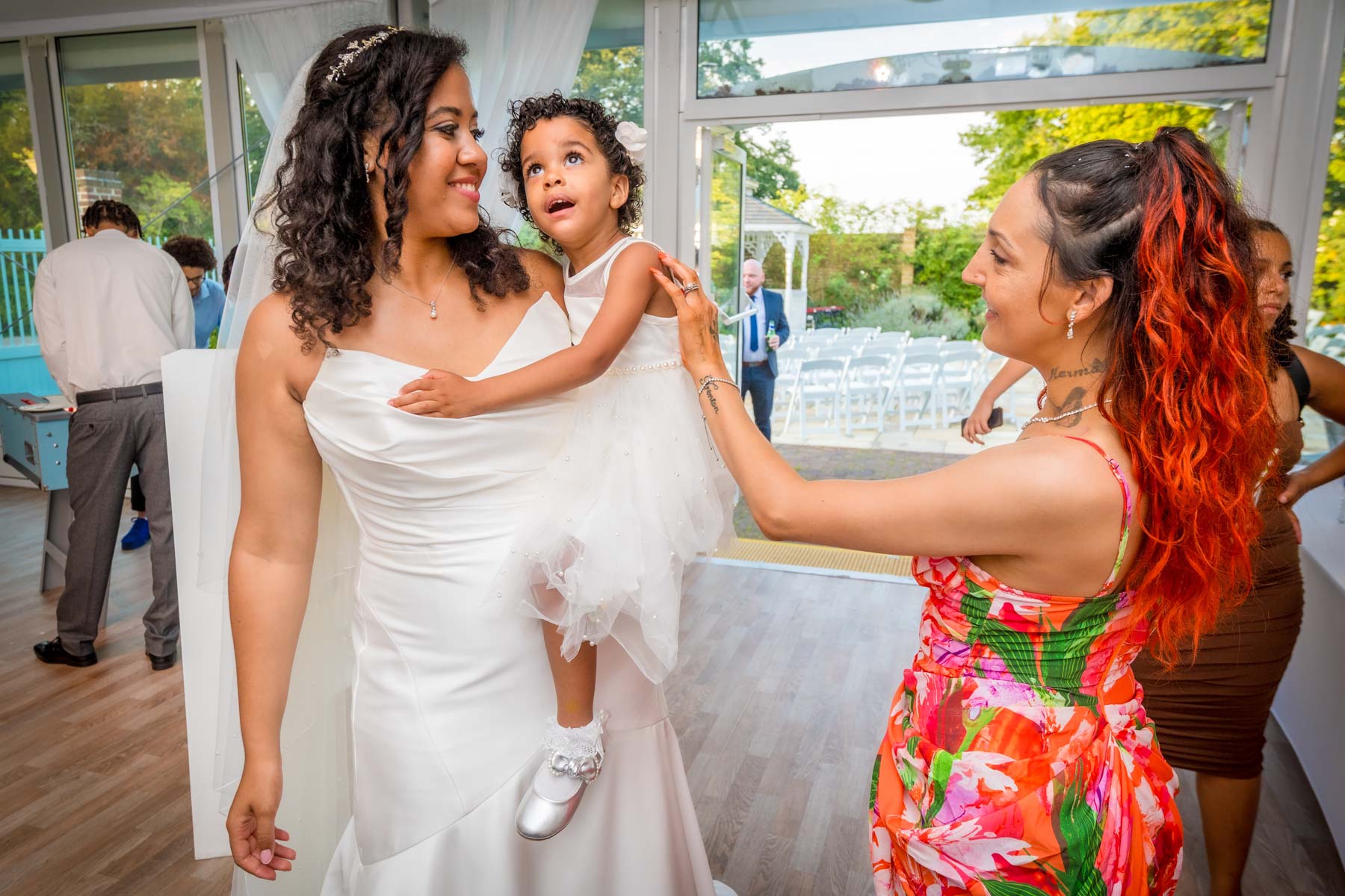 A natural photo of a bride carrying girl toddler whilst a guest touches her arm