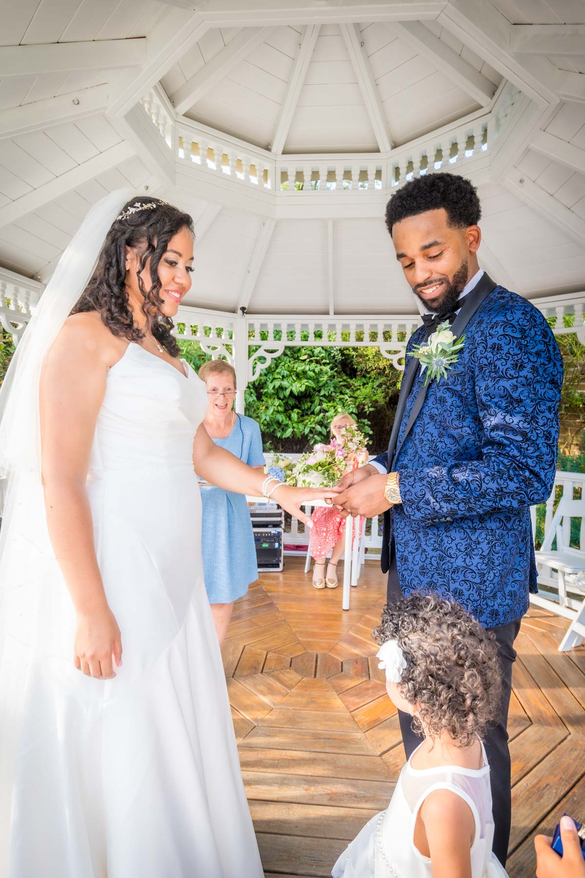 Groom places ring on bride's finger as little girl watches them at sunny wedding