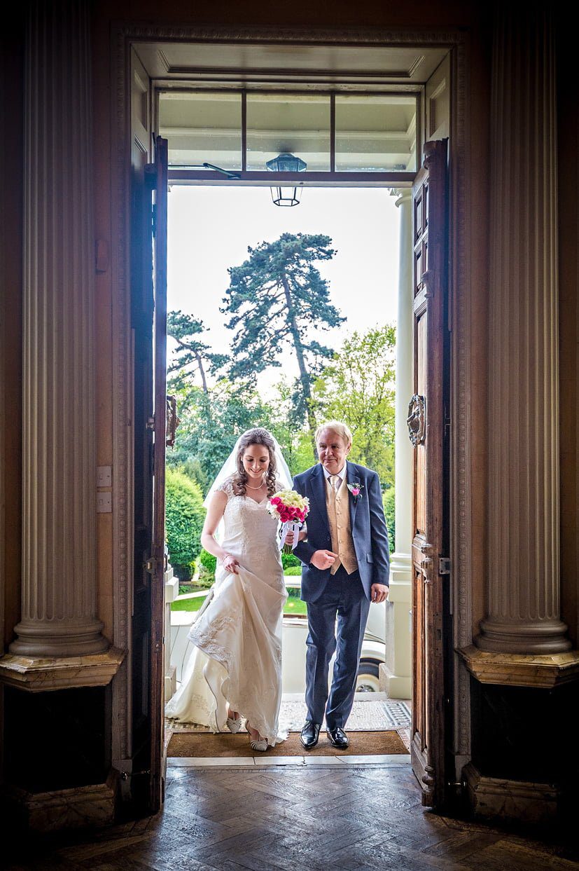 The bride arrives through the front doors of Hampton Court House with her father