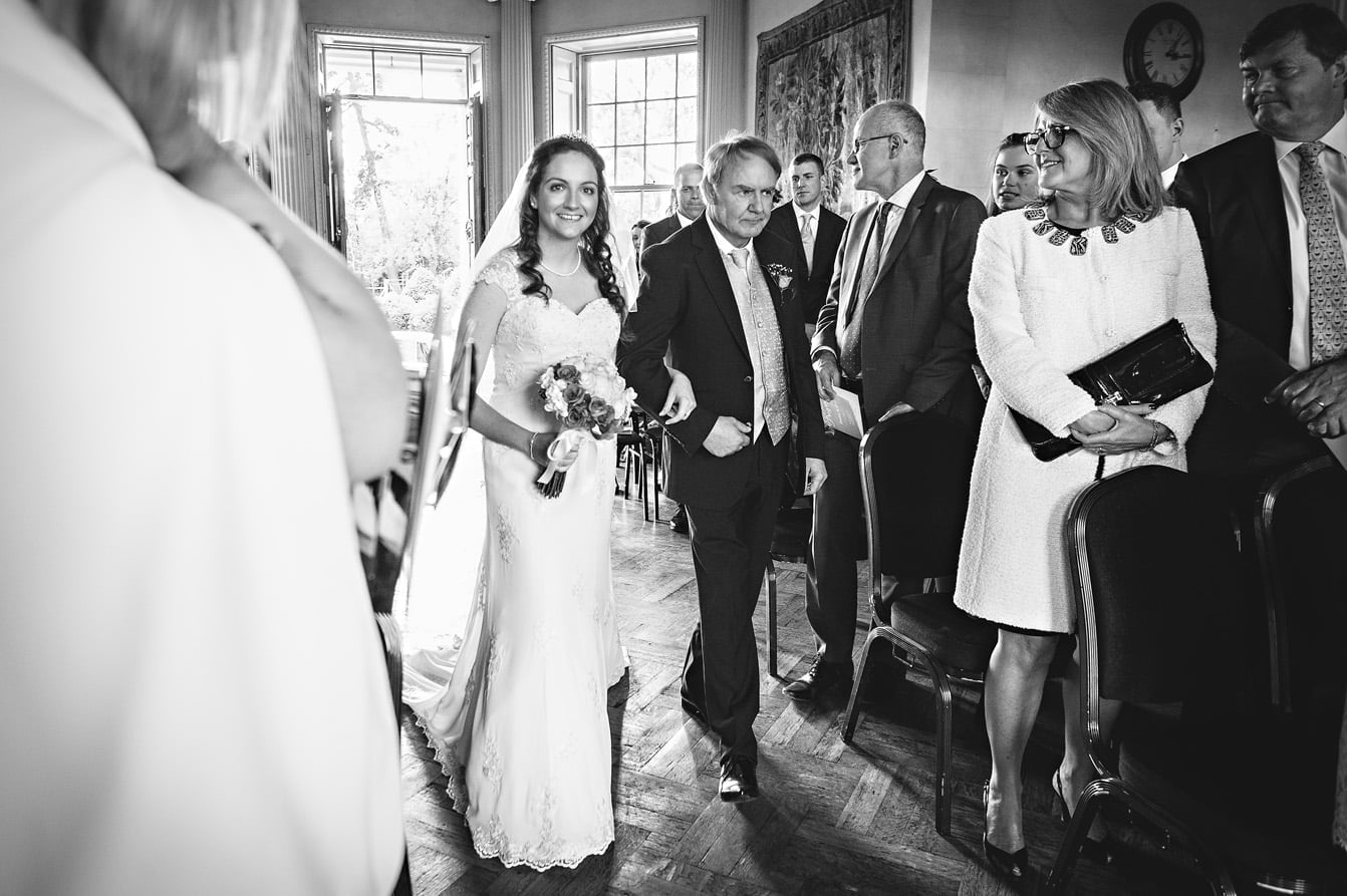 The bride walks down the aisle of the Main Hall in Hampton Court House with her father