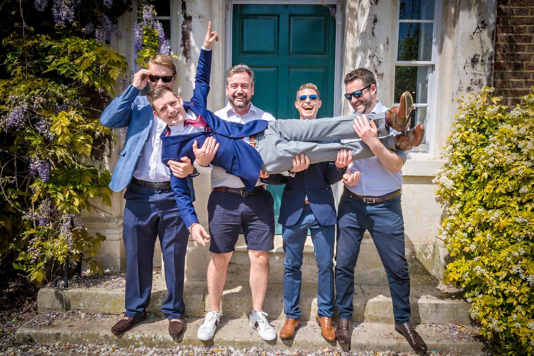 Four male wedding guests carrying groom outside front door in the sunshine