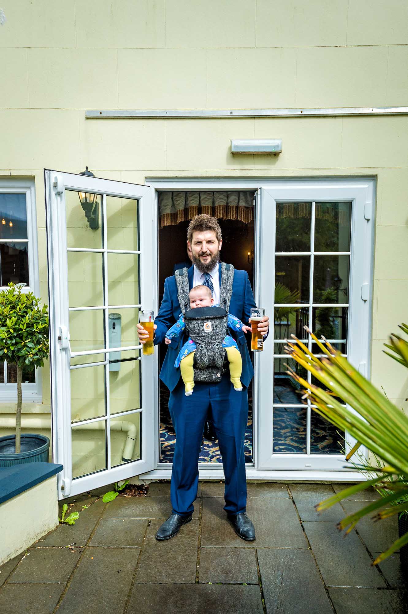 Bearded wedding guest with baby in carrier and two pints of beer leaving hotel