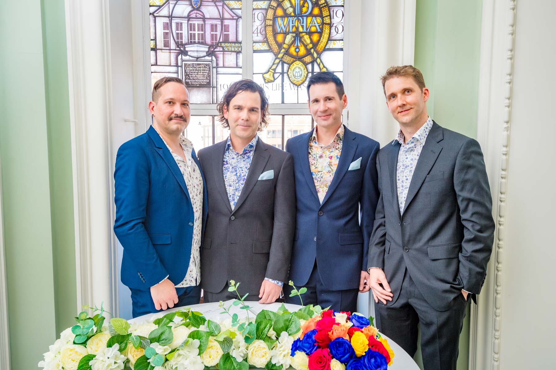 The grooms and witnesses pose behind a floral arrangement after their wedding in Brixton