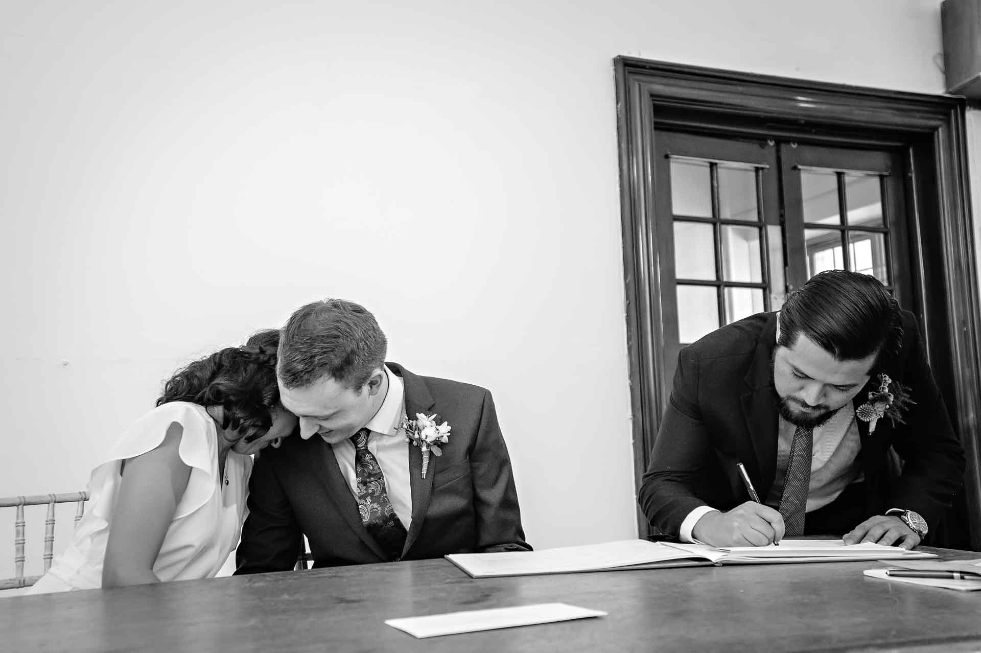 Bride nuzzles groom as witness signs the register