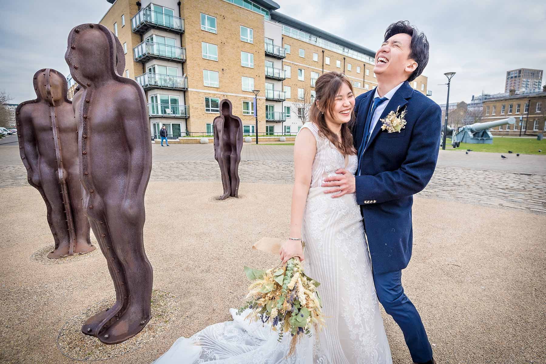 The wedding couple laughing as they pose with sratues at the Royal Arsenal Heritage Site in Woolwich, London