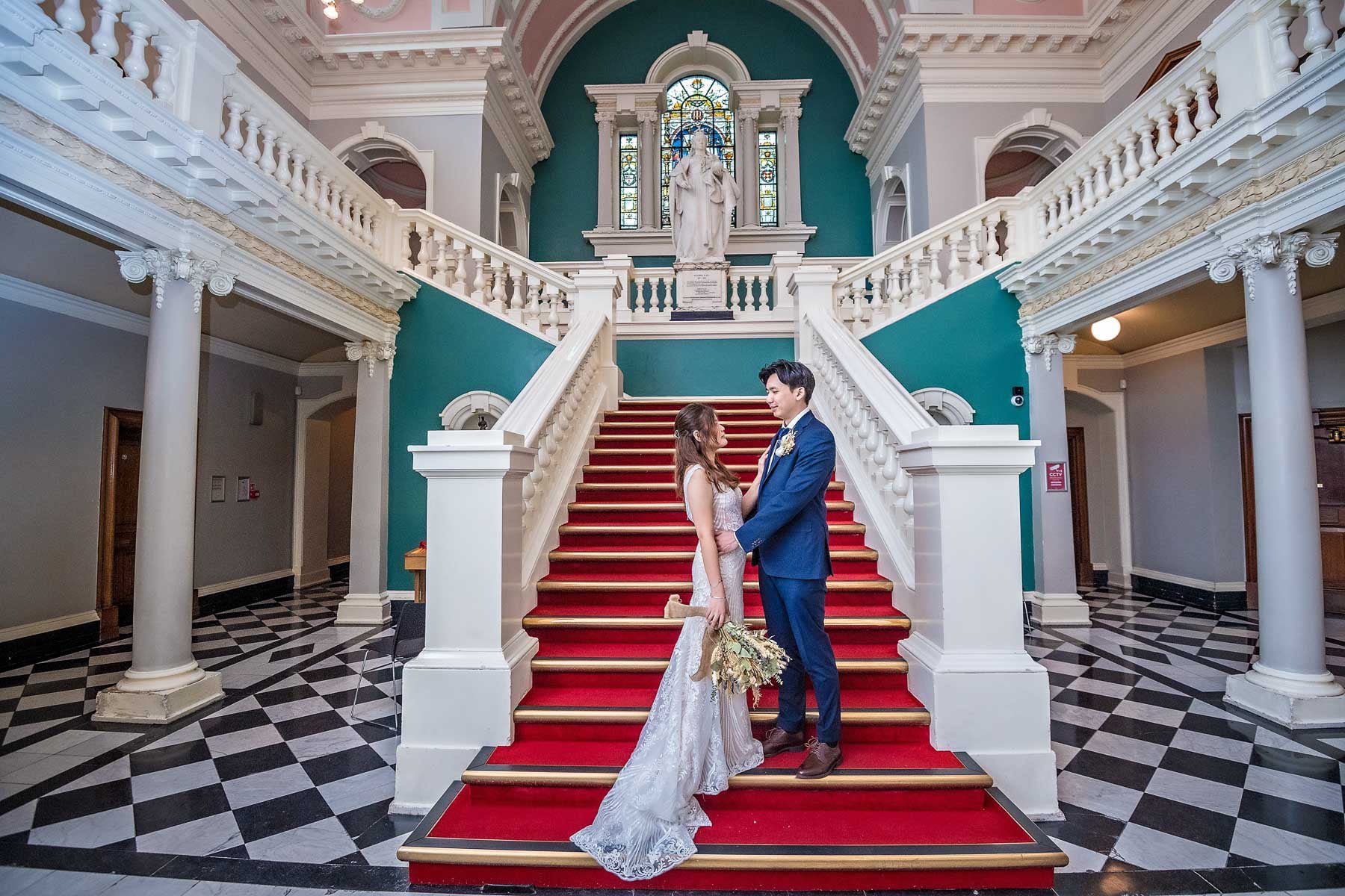 The bride and groom standing facing each other on the stairs of Vicoria Hall, Greenwich Town Hall, Woolwich