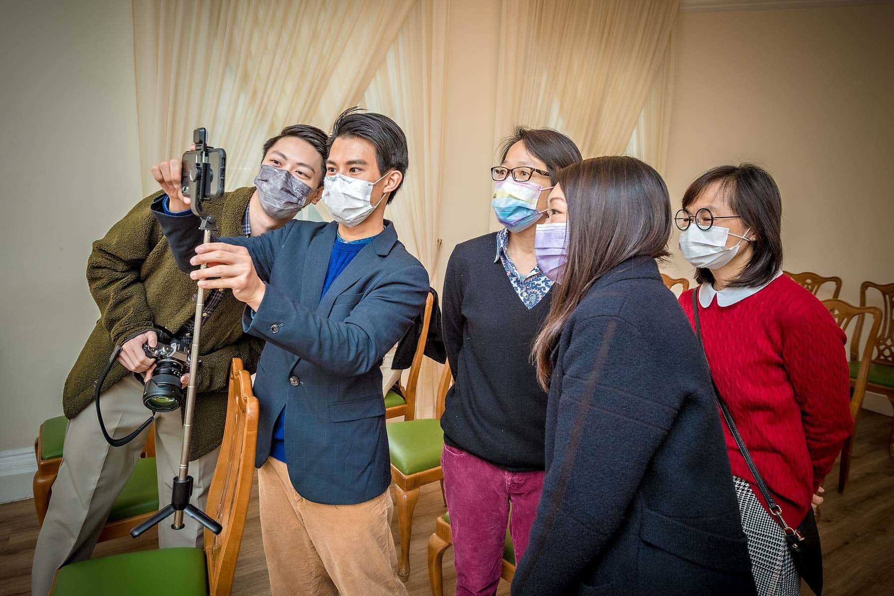 The wedding guests wearing Covid masks chat to relatives on a phone with selfie stick