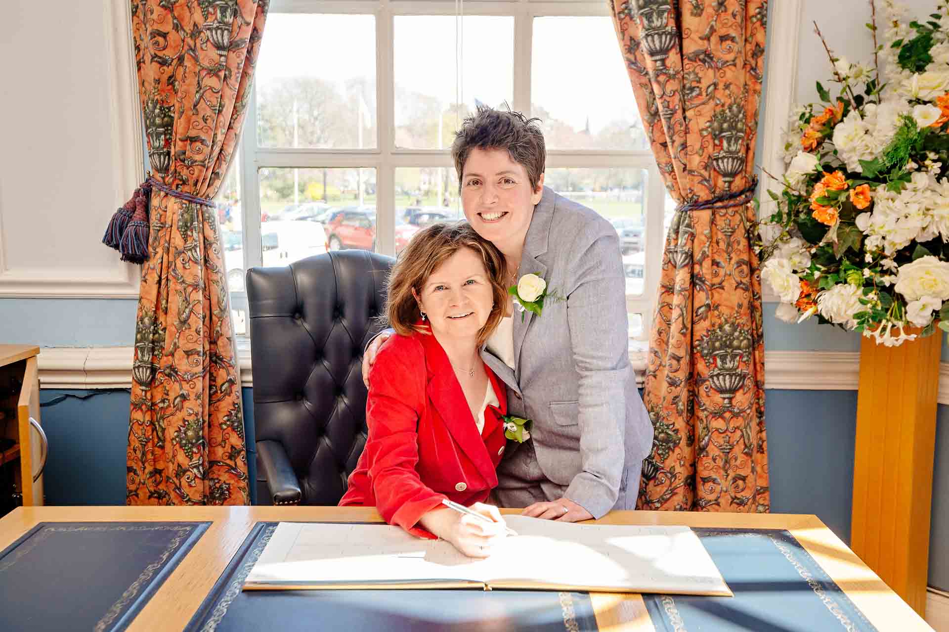 Brides posing for signing of register at desk in front of window