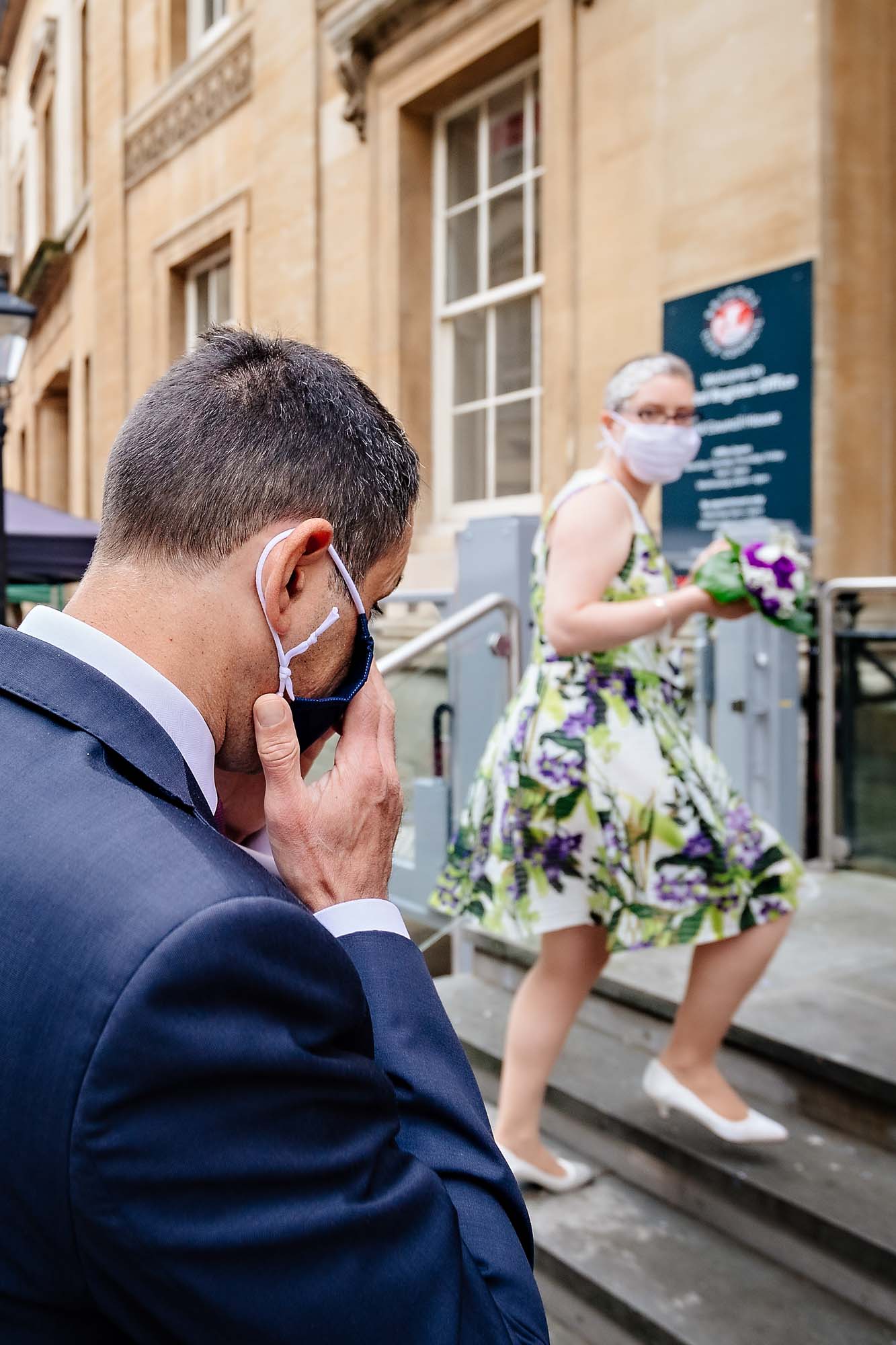 Groom puts on mask whilst entering wedding venue with bride watching him