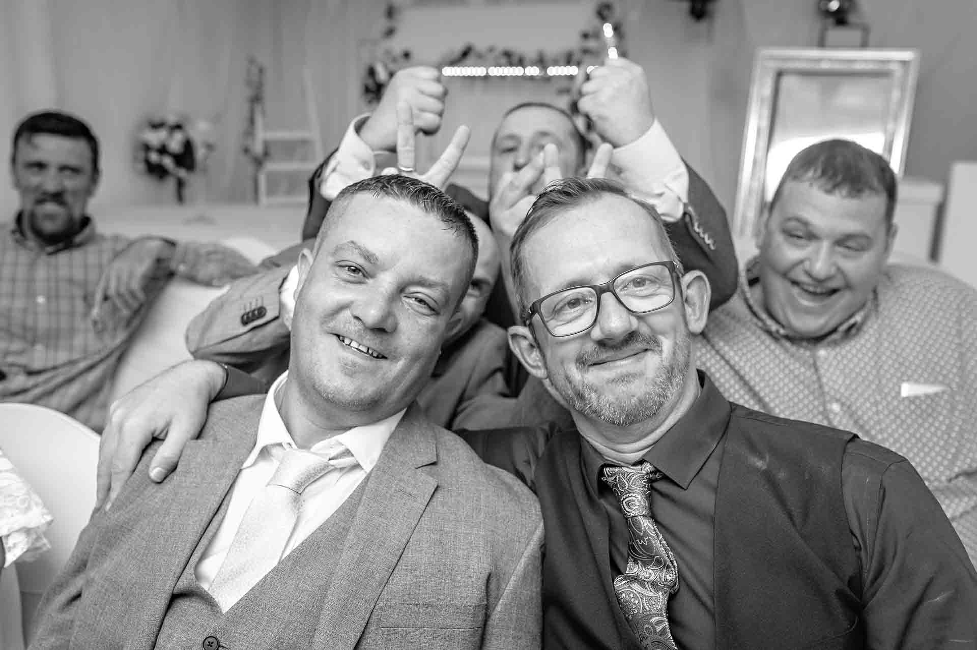 Two males friends pose whilst friend photo bomb background gesturing