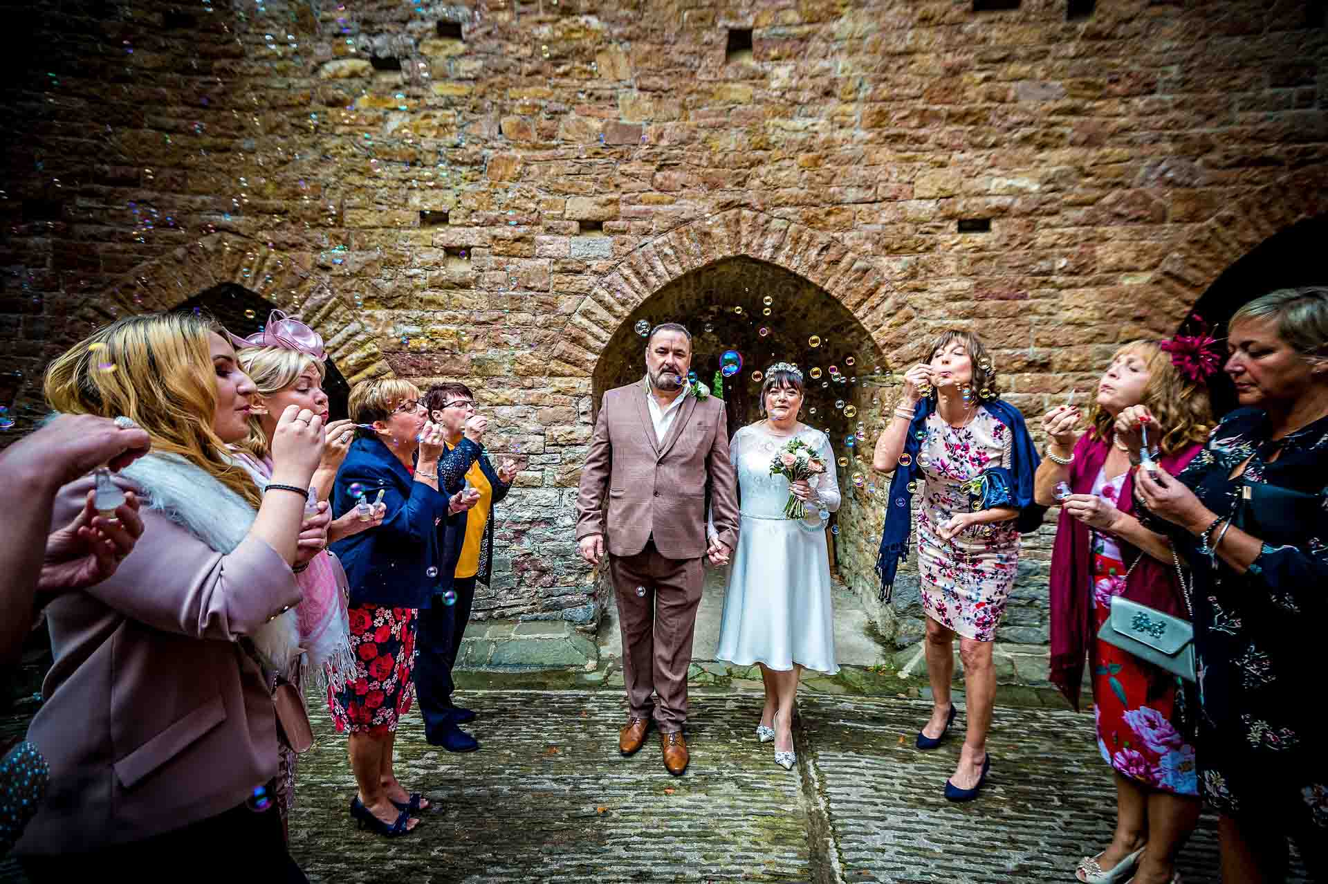 Wedding guests blowing bubbles as couple watch framed by an alcove's arch