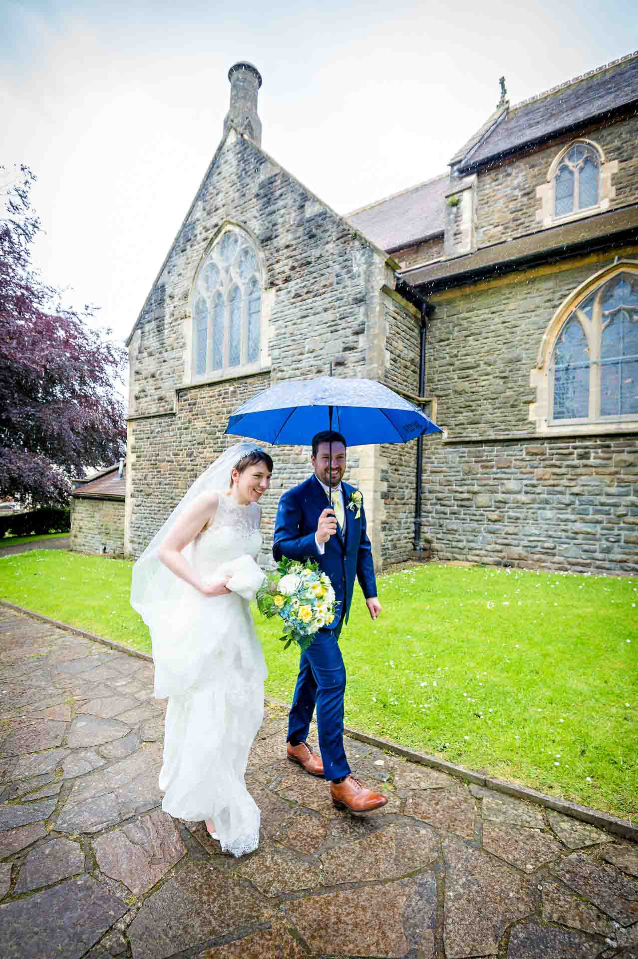 Bride and groom walking outside church in rain with umbrella
