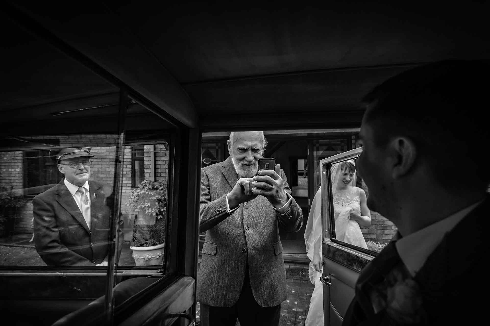 Bride's grandfather taking photo with phone of guests in car with bride watching