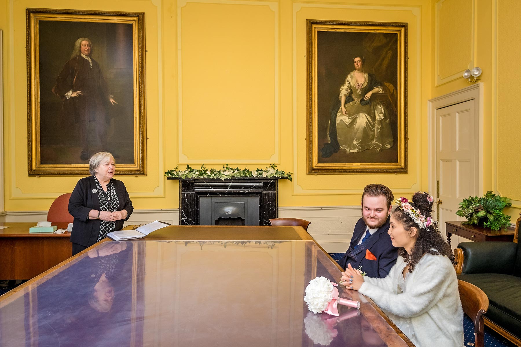 In the new weddings statutory room at Bristol Register Office with long table and portraits, the groom looks at his bride whilst the registrar watches on.
