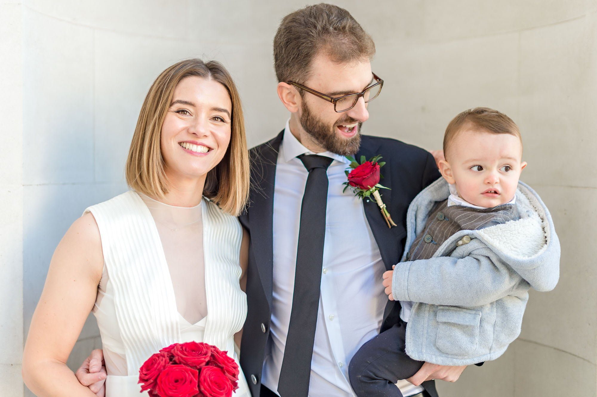 Bride and groom pose with little boy and red bouquet at London wedding