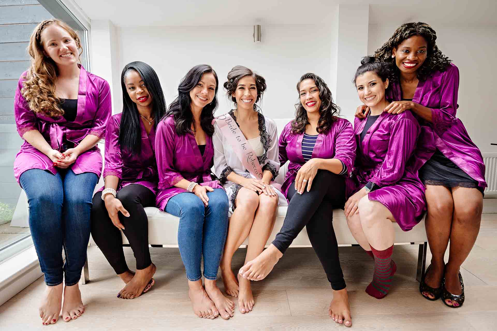 Bride seated with six bridesmaids in matching purple dressing gowns on couch