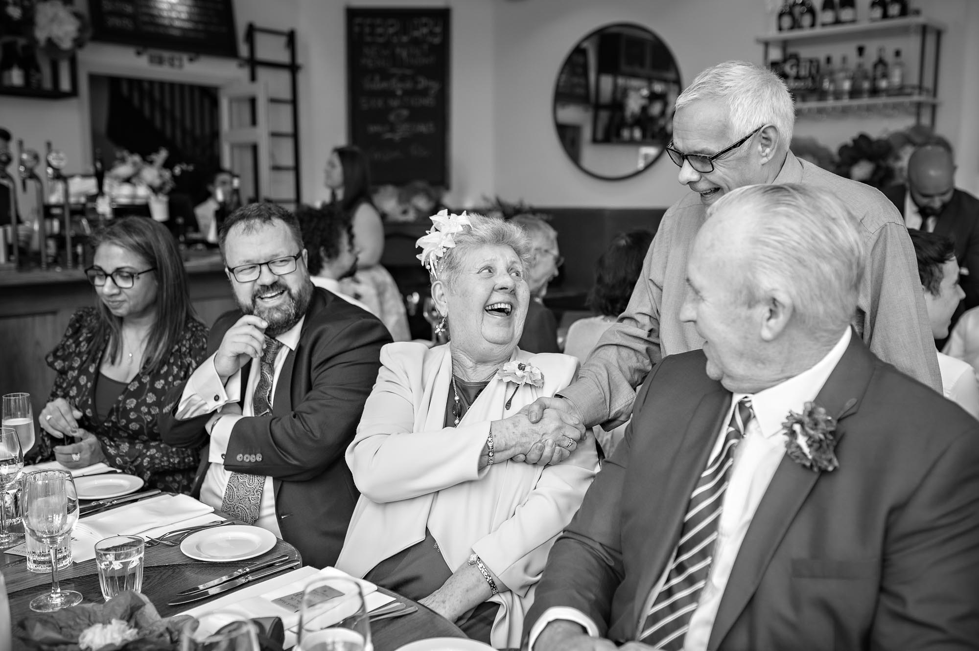 Mature lady shakes hand of man whilst sitting at table laid for meal with other guests watching