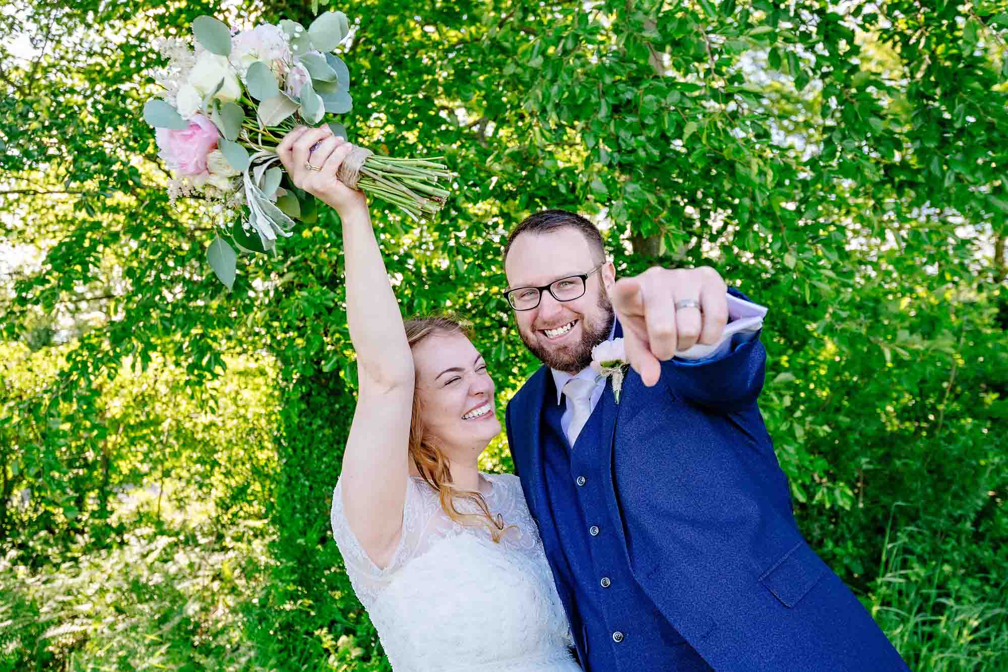 Portrait of bride and groom with groom pointing at the camera and bride holding bouquet in the air