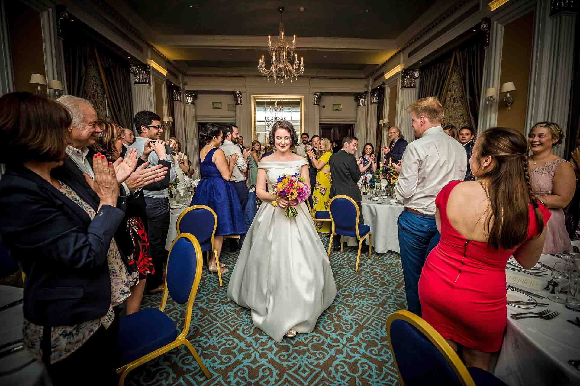 Bride walking through dining room whilst guests applaud at RAF Club wedding