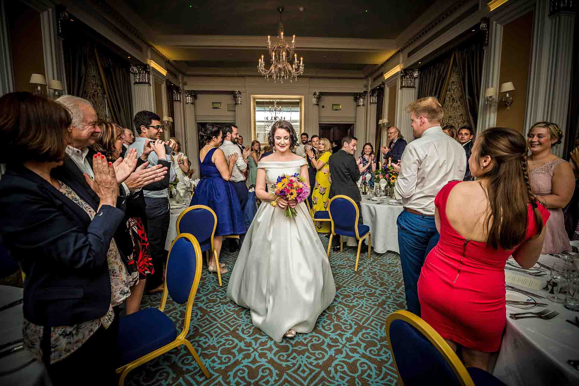 Bristol Wedding - Bride Enters the Wedding Breakfast Bride walking through dining room whilst guests applaud at RAF Club wedding