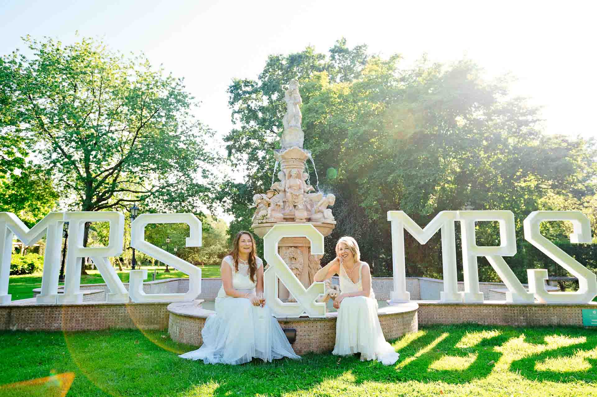 Two LGBTQ+ brides sitting in front of fountain with Mrs & Mrs sign