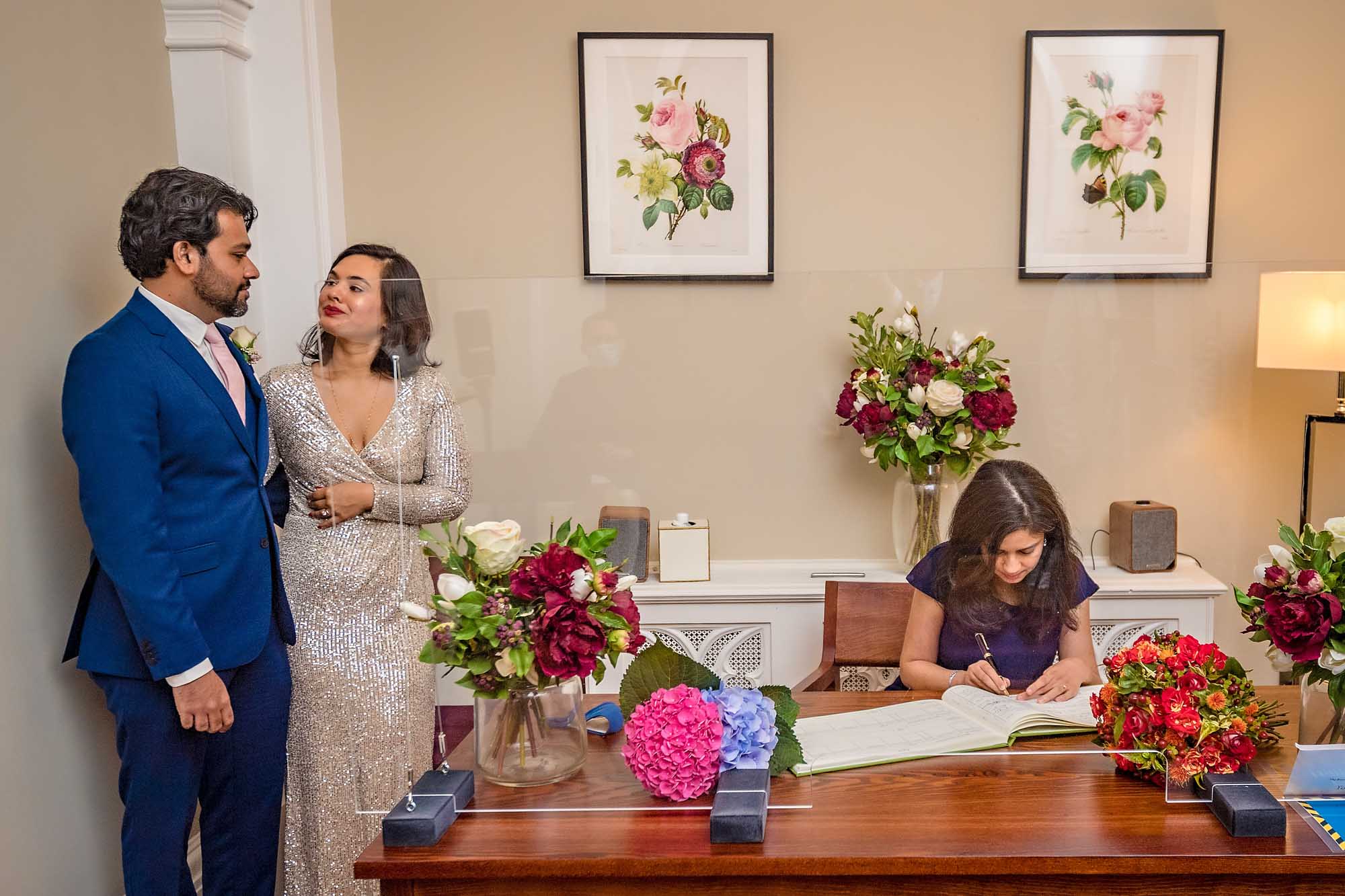 A witness signs the wedding register with couple behind in the Harrington Room, Chelsea Old Town Hall