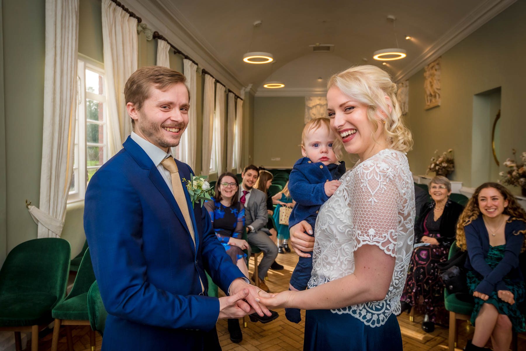 Exchange of rings in Loggia Room of York House, Twickenham. The couple smile as their baby son watches them.