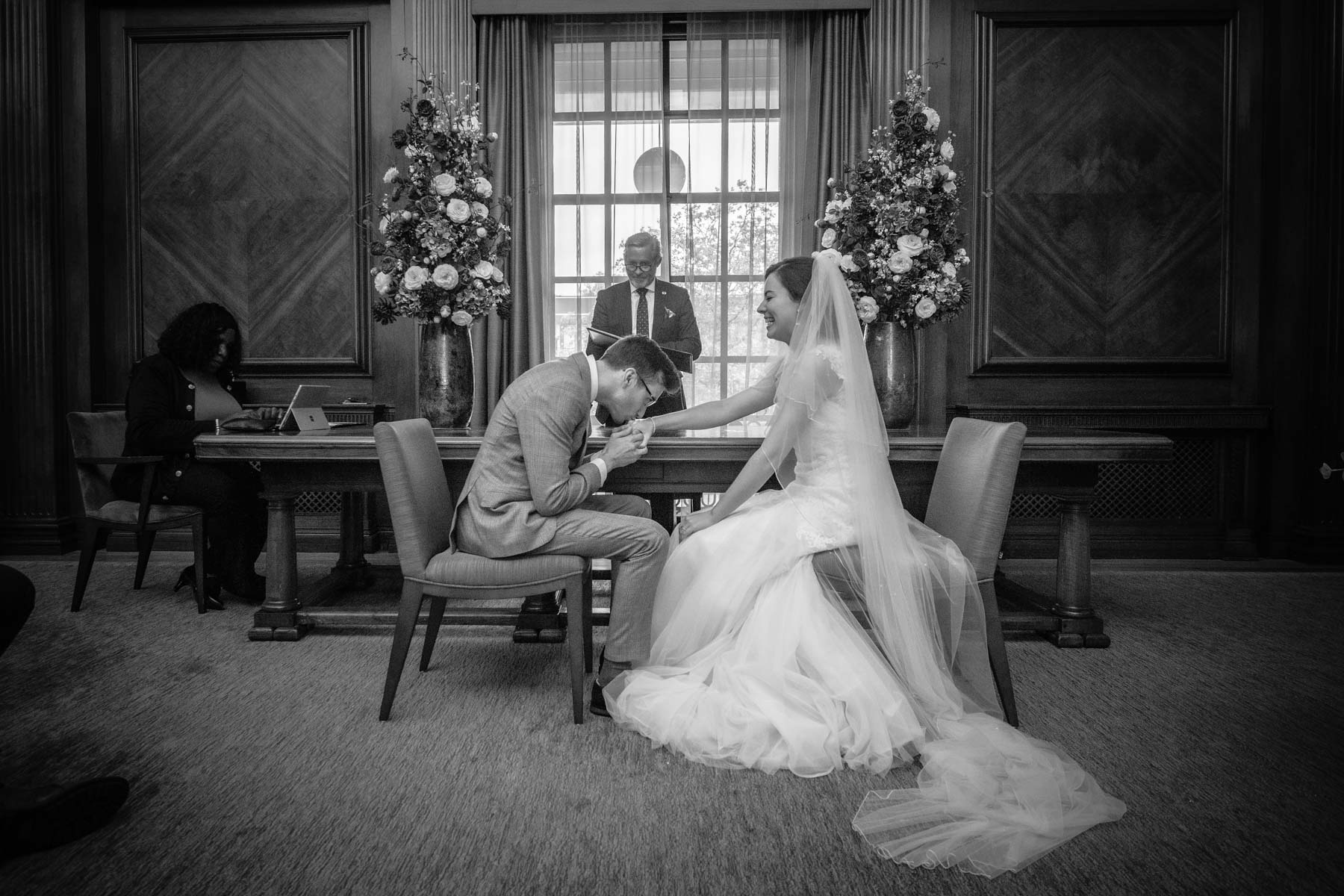 Groom kisses his bride's hand whilst she laughs as they're seated at their wedding.